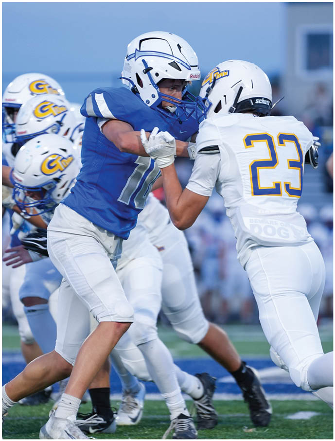 Two people in football uniforms are engaged in a blocking action on a field with other players visible in the background. Description generated by AI