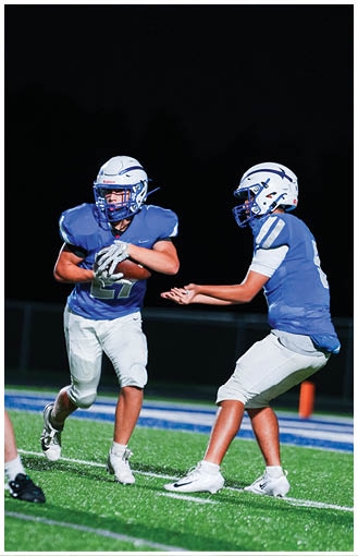A person wearing a blue football uniform and white helmet is running with a football while another person reaches out with both hands attempting to catch or tackle. Description generated by AI