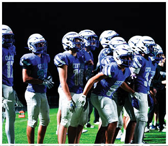 People wearing football helmets and uniforms stand together on a field under bright lights against a dark background. Description generated by AI