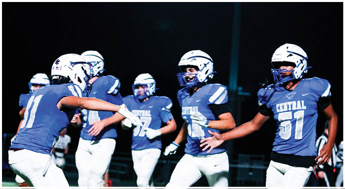Five people wearing football helmets and uniforms are on a field at night, appearing to be in the middle of a game or practice. Description generated by AI