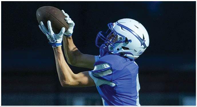 A person wearing a blue sports uniform and white helmet is catching a football during a game at dusk. Description generated by AI