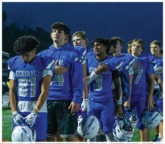 People wearing football helmets and uniforms stand together on a field under bright lights against a dark background. Description generated by AI