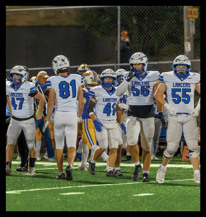 People wearing football uniforms are standing on a field during a game or practice, with some facing away and others facing forward. Description generated by AI