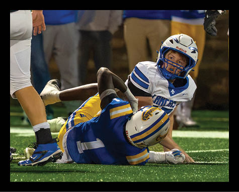 Two football players in helmets and uniforms are on the ground during a game, with one player in white looking up while the other in blue and yellow is lying down. Description generated by AI