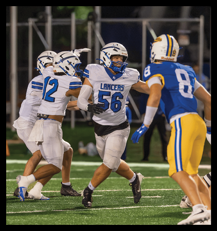 A person wearing a white football uniform with the number 56 and the word Lancers is running on a field while another person with a blue glove reaches out. Description generated by AI