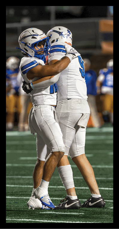 Two football players in white uniforms and helmets are embracing on a field during a game. Description generated by AI