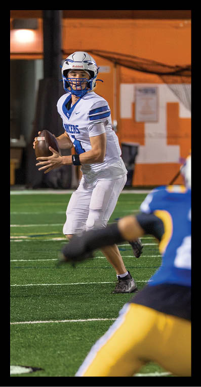 A person in a white and blue football uniform is preparing to throw a football while another person in a blue and yellow uniform is moving toward them on a football field. Description generated by AI