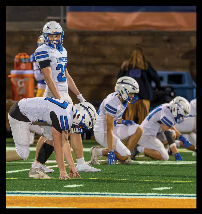 Three people in white football uniforms and helmets are crouched on a green field, preparing for a play. Description generated by AI