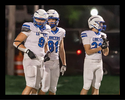 Three people wearing white and blue football uniforms with helmets stand on a field at night. Description generated by AI