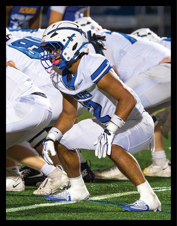 A person in white football gear and helmet is crouched in a ready position on a green field with other players in similar uniforms lined up behind. Description generated by AI