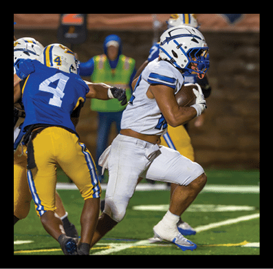A person in a white football uniform is running with the ball while another person in a blue and yellow uniform reaches out to tackle them on a football field. Description generated by AI