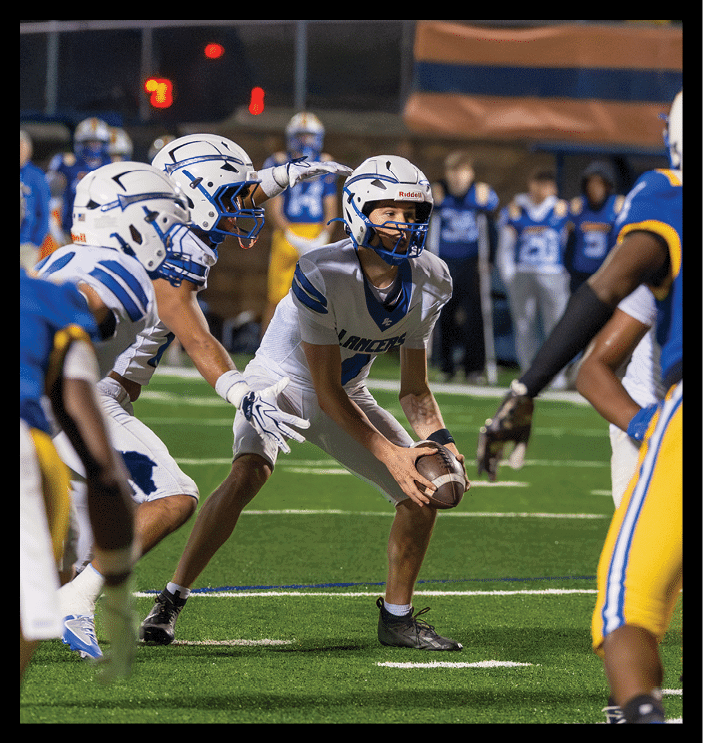 A person in a white football uniform and helmet is holding a football ready to snap it while another person in a similar uniform reaches out toward them on a football field during a game. Description generated by AI