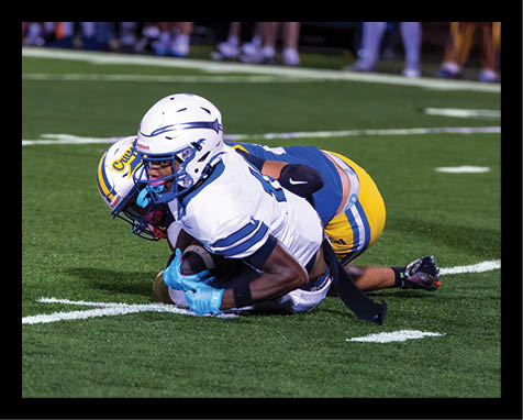 Two people in football uniforms are on the ground during a tackle on a grassy field. Description generated by AI