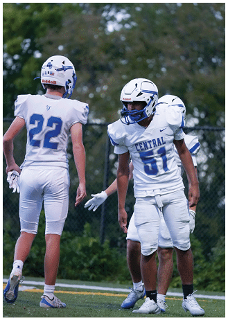 Three people in white football uniforms and helmets stand on a field with trees and a fence in the background. Description generated by AI