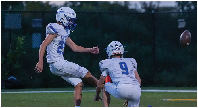 A person wearing a white and blue football helmet and jersey is on a field with trees and a tower visible in the background at dusk. Description generated by AI