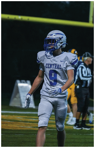 A person in a football uniform with the number nine and the word Central on the jersey is walking on a football field near a goalpost during a game. Description generated by AI