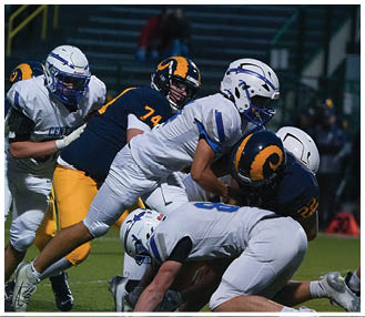 Two people wearing football helmets and uniforms are engaged in a play during a game, with one person in white leaning forward and the other in dark blue and yellow behind them. Description generated by AI