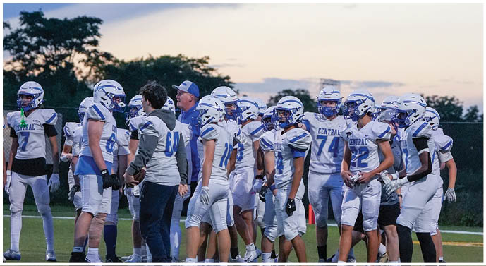 A group of people wearing football helmets and uniforms are gathered outdoors on a field with trees and a cloudy sky in the background. Description generated by AI