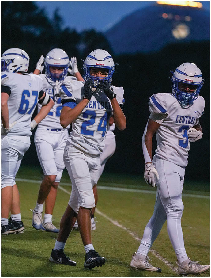Four people in white football uniforms with blue helmets stand on a field at dusk with a mountain and illuminated structure in the background. Description generated by AI