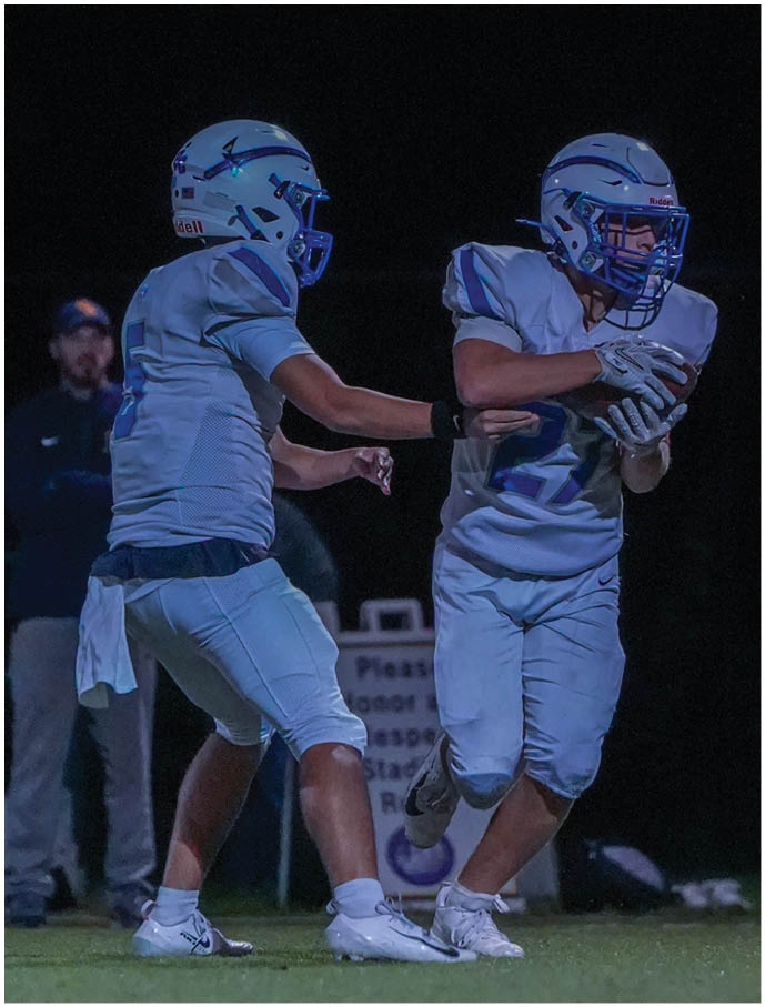 A person in a white football uniform and helmet is preparing to hand off the ball to a teammate during a nighttime game. Description generated by AI