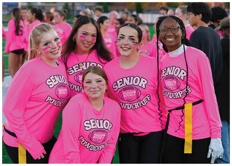 A person with long hair and pink face paint is standing next to a bright pink shirt with the word “SENI" and a football graphic partially visible. Description generated by AI