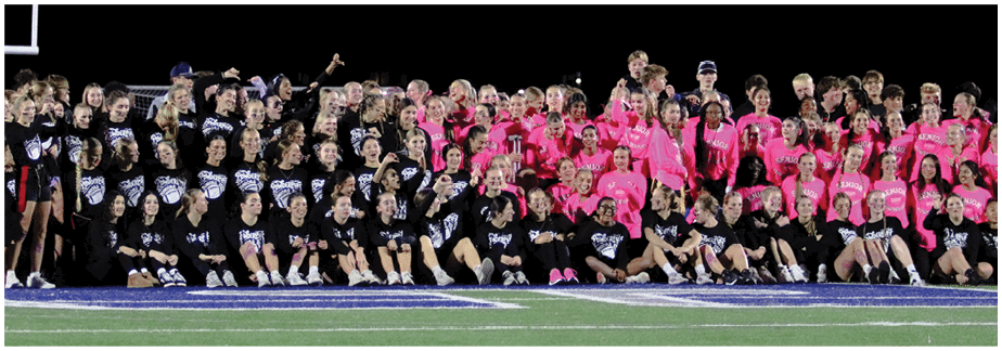 A large group of people wearing matching black and pink shirts are gathered closely together, smiling and interacting in a lively outdoor setting. Description generated by AI