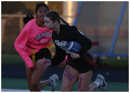 Two people are playing flag football, with one person holding a blue and white football while the other wears a bright pink shirt with “SENIOR 2027" printed on it. Description generated by AI