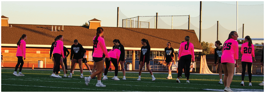 People wearing bright pink shirts are gathered outdoors near a brown building with a fenced area and clear sky in the background. Description generated by AI