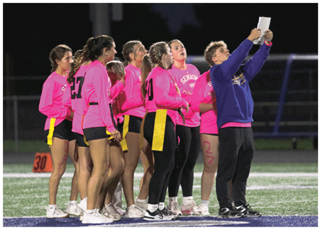 A group of people wearing bright pink shirts and yellow flags stand on a sports field while one person in a blue sweatshirt holds up two pieces of paper. Description generated by AI