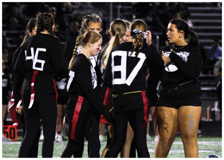 People wearing black athletic clothing with red flags are standing on a field, some with numbers on their backs and markings on their legs. Description generated by AI