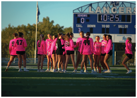 A group of people wearing bright pink sports jerseys and shorts are standing on a grassy field with a fence and trees in the background. Description generated by AI