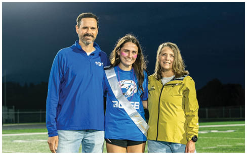 Three people stand together on a sports field at night with one person wearing a sash and a blue shirt, flanked by two others in casual jackets and jeans. Description generated by AI
