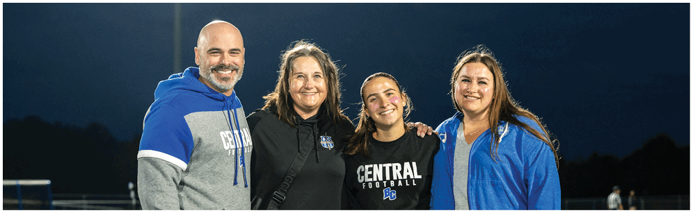 Four people standing closely together on a sports field at night, smiling and wearing casual and athletic clothing including hoodies and a shirt with “Central Football" printed on it. Description generated by AI