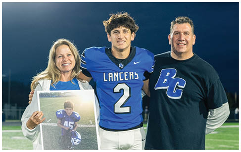 Three people stand on a football field at night with one person in a blue and white football uniform holding a framed photo of a person in a similar uniform. Description generated by AI