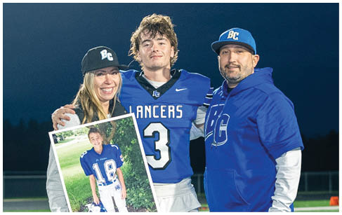 Three people stand on a football field at night with one person in a football uniform holding a framed photo of a child in a football uniform. Description generated by AI