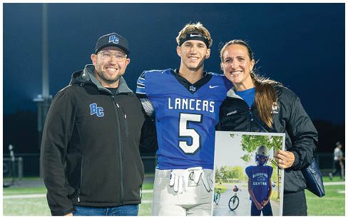 Three people stand on a football field at night with one person in a blue and white football uniform and the other two in casual jackets, while one holds a framed photo of a child. Description generated by AI