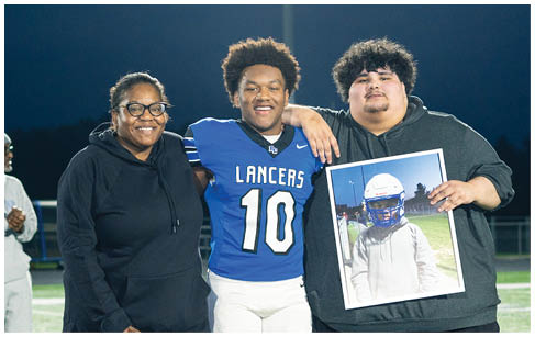 Three people stand on a football field at night with one person in a blue football uniform smiling between two others, one holding a framed photo of a person in football gear. Description generated by AI