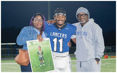 A person in a football uniform stands on a field at night with two people, one holding a photo of a child in a sports uniform, all smiling and posing together. Description generated by AI