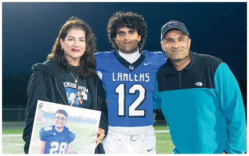 A person in a football uniform stands on a field at night between two people holding a framed photo of a person in a football uniform with a child. Description generated by AI
