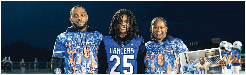 Three people stand on a football field at night wearing Lancers-themed clothing, with one person holding a photo of a child in a football uniform. Description generated by AI