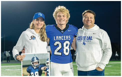 Three people stand on a football field at night with one person in a blue football uniform and the other two in white hoodies, one holding a framed photo of a football player. Description generated by AI