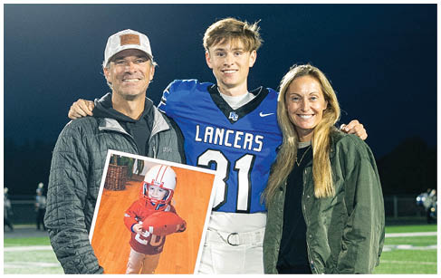 A person in a football uniform stands on a field at night with two adults, one holding a framed photo of a child in a football outfit. Description generated by AI