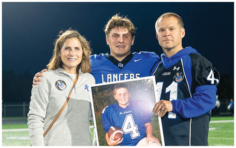 Three people stand on a football field at night with one person in a football uniform holding a framed photo of another person in a football uniform. Description generated by AI