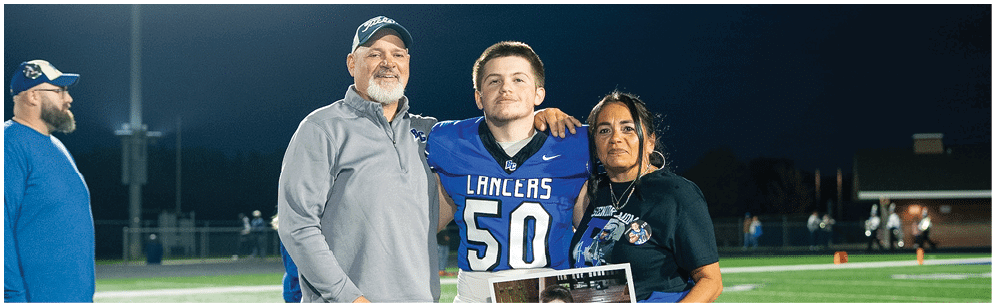 Three people stand on a football field at night with one person in a football uniform holding a framed photo of another person in a football uniform. Description generated by AI