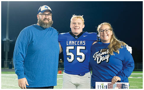 Three adults stand on a football field at night with one wearing a football uniform and the other holding a photo of a child in a football uniform. Description generated by AI