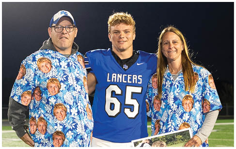 Three people stand on a football field at night with two wearing matching blue floral shirts printed with a person's face and the third wearing a blue football uniform holding a large photo of a child in a football uniform. Description generated by AI
