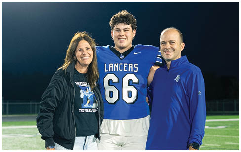A person in a football uniform stands on a field at night with two adults, one holding a framed photo, all smiling under bright stadium lights. Description generated by AI