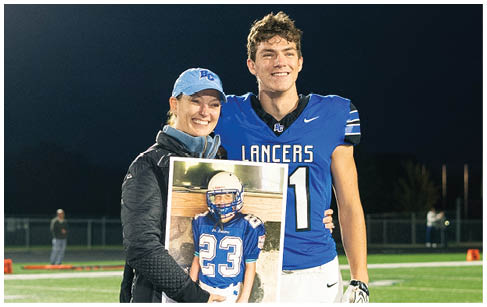 A person in a blue sports jacket and cap stands on a football field at night holding a framed photo of another person in a blue football uniform while standing next to a person in a blue football jersey and white pants. Description generated by AI