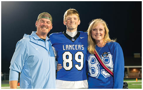 Three people stand together on a sports field at night with one person wearing a football uniform and the other two wearing casual clothing with matching blue tops. Description generated by AI