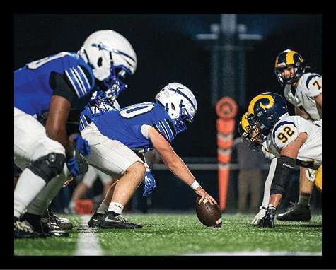 A person in a blue football uniform is crouched down on a field, holding a football on the ground, preparing for a play. Description generated by AI